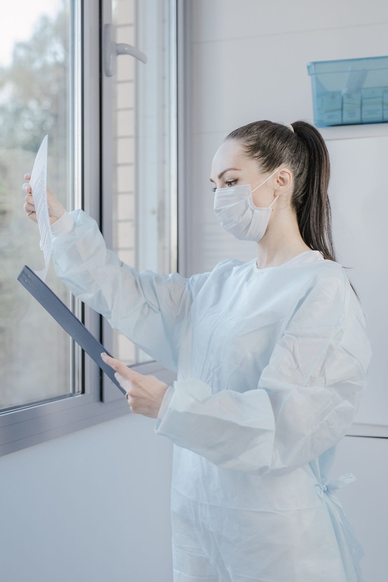 Clinician in surgical gown and mask reviewing a clipboard document by a bright clinical window