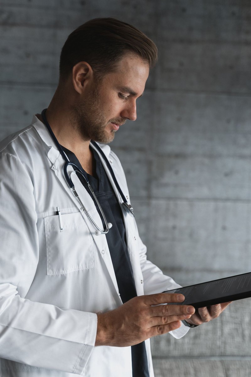 Male doctor in white coat with stethoscope reviewing a black tablet against a concrete wall